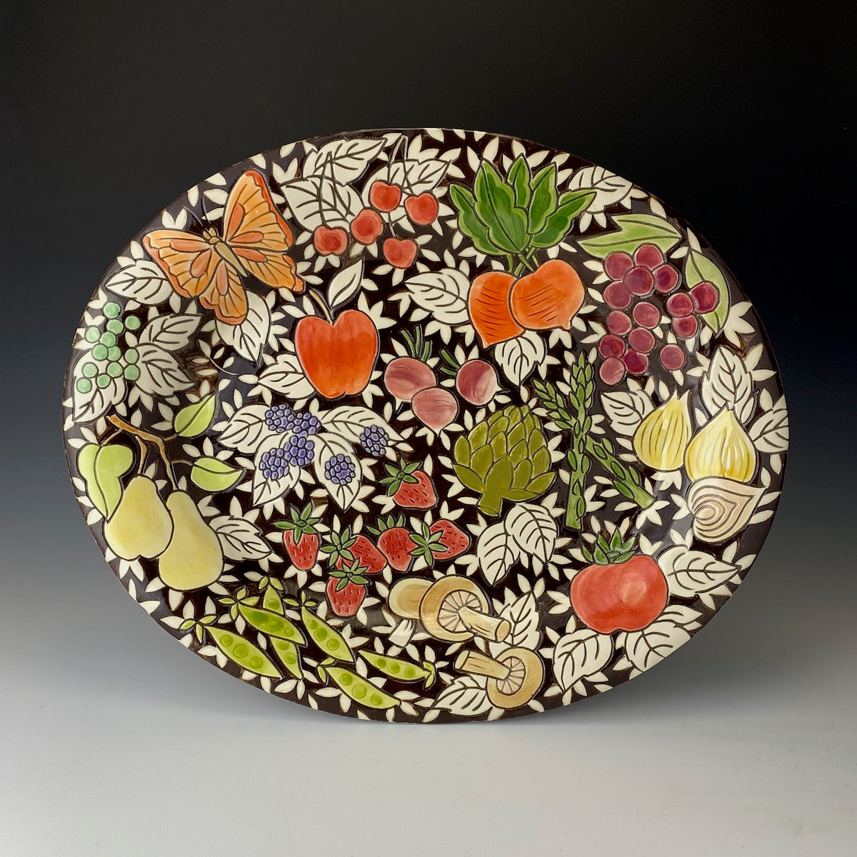 Decorative ceramic platter with colorful fruit, vegetable, and butterfly design on a dark background